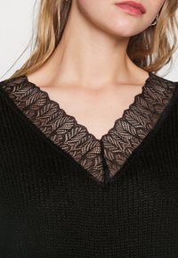 Femme portant un haut tricoté noir avec un décolleté en V orné d'une bordure en dentelle noire avec des motifs floraux.