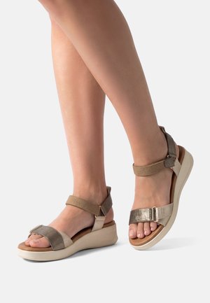 Piernas femeninas que llevan sandalias beige y metálicas con punta abierta, con correas en el tobillo y en los dedos, apoyadas sobre una superficie clara.