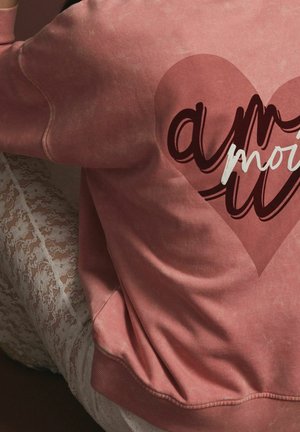 Sweat-shirt rose avec un grand graphique de cœur, accompagné de texte courbé en blanc et bordeaux. Tissu doux et texturé avec des poignets côtelés.