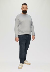 Grauer Langarm-Pullover, dunkelblaue Jeans, beigefarbene Sneakers mit hellen Akzenten. Das Model steht aufrecht vor einem neutralen Hintergrund.