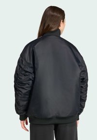 Une personne avec de longs cheveux bruns porte une veste bomber noire, vue de dos, sur un fond vert pâle.