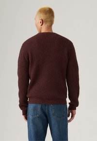 Man with short blonde hair wearing a textured dark burgundy sweater and blue jeans facing away against a plain background.