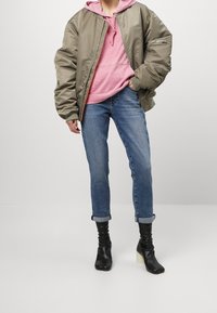 Chaqueta bomber verde oliva, sudadera rosa, jeans azules desgastados, botas negras de tobillo con tacones cuadrados. Conjunto casual en capas.