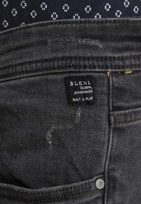 Close-up of black denim jeans pocket with a black fabric label reading "BLEND GLOBAL JEANSMAKER BUILT IN BLUE" and dark patterned fabric above.