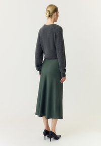 Kvinde med blond hår i knold iført en mørkegrå fluffy sweater, mørkegrøn satin midi nederdel og sorte høje hæle, vendt væk.