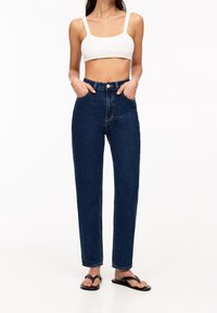 Vaqueros de mezclilla de tiro alto en azul oscuro con un corte recto, que cuentan con bolsillos frontales y cierre de botón, combinados con un top corto blanco y sandalias negras.