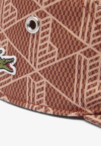 Cappello marrone con motivo, realizzato in tessuto testurizzato, dotato di un occhio metallico e di un logo coccodrillo ricamato in verde. Sono presenti motivi esagonali e diagonali.