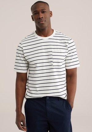 Mees, kes kannab valget lühikese varrukaga särki mustade horisontaalsete triipude ja rinnataskuga, tumeda mereväe pükstega, seisab tavalise tausta ees.