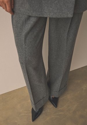 Pantalon gris à jambes larges avec revers porté avec des chaussures noires à bout pointu sur un sol marron près d'un mur beige.