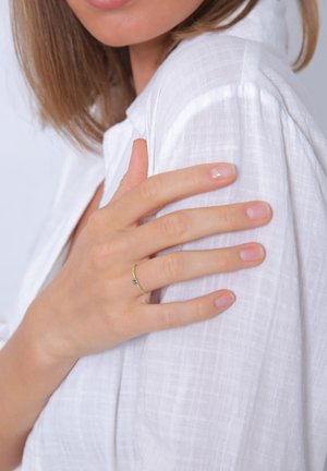 Goldring mit einem kleinen blauen Edelstein, der an einer Hand getragen wird. Die Hand liegt auf einem weichen, weißen, strukturierten Stoff. Helle, neutrale Nagellack.