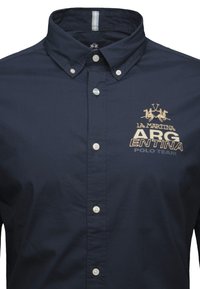 Camisa de botones azul marino hecha de algodón con cuello clásico, que presenta un logo y el texto "ARGENTINA POLO TEAM" en dorado. Textura sólida.