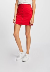 Frau in einem roten Minirock mit Knopfleiste und frontalen Taschen, kombiniert mit weißen High-Top-Sneakern, steht vor einem schlichten Hintergrund.