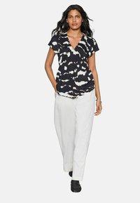 Blusa de tie-dye negra y crema con mangas cortas, escote en V y detalles fruncidos, combinada con pantalones anchos blancos y zapatos negros.
