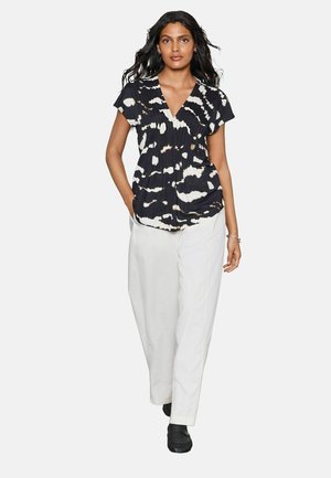 Schwarze und cremefarbene Batikbluse mit kurzen Ärmeln, V-Ausschnitt und gerafften Details, kombiniert mit weißen weit geschnittenen Hosen und schwarzen Schuhen.
