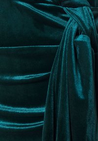 Tissu en velours de couleur vert profond, avec des détails froncés et un accent noué. Texture lisse avec un léger éclat, mettant en valeur une couleur riche.