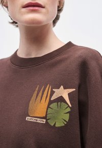 Καφέ φούτερ με εκτύπωση γραφικού στοιχείου που απεικονίζει κίτρινα-πορτοκαλί φύλλα, ένα καφέ αστέρι και ένα πράσινο διακοσμητικό στοιχείο, με την ένδειξη "SCOTCH & SODA".