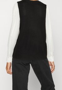 Top nero senza maniche con scollatura rotonda, abbinato a una camicia bianca a maniche lunghe con pois sotto. I jeans scuri completano l'outfit.