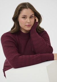 Bordeauxfarbener Strickpullover mit gerippter Textur an den Ärmeln und Stehkragen; glattes Material am Körper; getragen mit einer Hand, die auf einem weißen geometrischen Objekt ruht.
