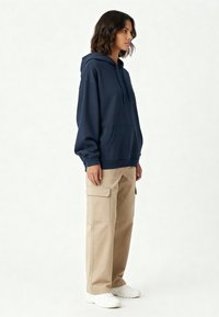 Felpa blu navy con tasca a marsupio e cordoncino regolabile, abbinata a pantaloni cargo beige con tasche laterali e sneakers bianche.