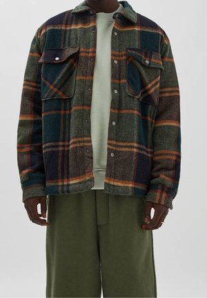 Mann trägt eine grün-braun-orange karierte Jacke mit Knöpfen über einem hellgrünen Sweatshirt und olivgrünen Hosen und steht mit entspannten Händen.