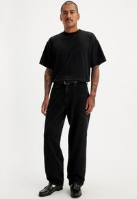 Moški s tetovažami, oblečen v črno preveliko T-majico, ohlapne črne kavbojke, črni pas in črne loafer čevlje, stoji ob belem ozadju.