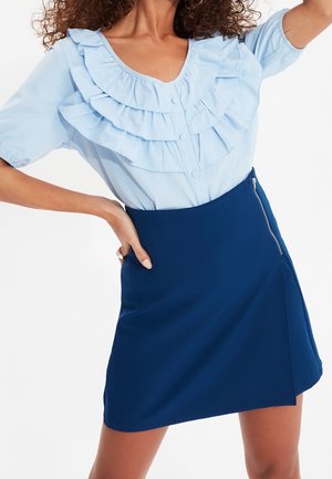Frau trägt eine hellblaue, mit Rüschen versehene Bluse, die in einen dunkelblauen A-Linien-Rock mit sichtbar silbernem seitlichem Reißverschluss gesteckt ist.