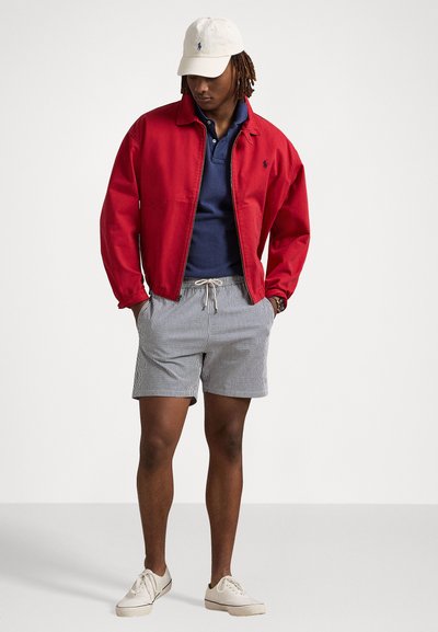 Red bomber jacket, navy polo shirt, striped shorts, cream sneakers, and a light cap with a logo. Casual outfit with varied textures and colors.