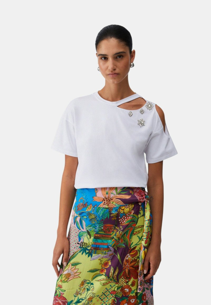 Woman wearing a white short-sleeve top with shoulder cutouts and jeweled embellishments, paired with a colorful floral patterned skirt.