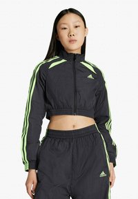 Junge Frau trägt schwarze und neongrüne Adidas-Cropped-Jacke und passende Hose, steht vor einem schlichten weißen Hintergrund.
