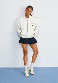 Jaqueta bomber branca com um bolso com fecho, usada sobre uma camisa cinza, combinada com uma saia preta e ténis com riscas verdes num chão azul.