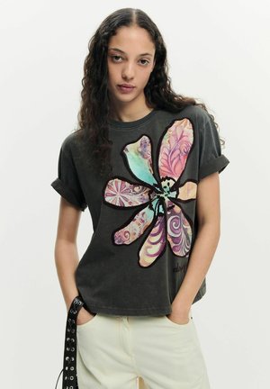 Jeune femme aux cheveux longs et bouclés portant un T-shirt noir avec un grand motif floral coloré et un pantalon clair.