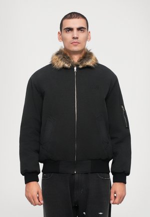 Schwarze Bomberjacke mit einem Kragen aus Kunstfell, Reißverschluss, seitlichen Taschen, gerippten Bündchen und Saum. Detaillierte Nähte in der Nähe der Brust.