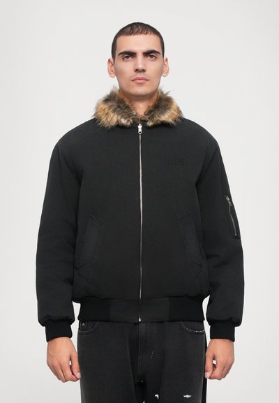 Veste bomber noire avec col en fausse fourrure, fermeture éclair, poches latérales, poignets et ourlet côtelés. Couture détaillée près de la poitrine.