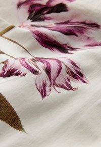 Tyg med ett blommönster med lila och rosa stiliserade blommor och bruna blad på en ljusbeige bakgrund.
