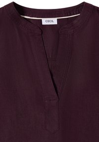 Chemise violet foncé avec un col rond et une encoche en forme de V, avec des coutures visibles et une étiquette blanche portant l'inscription « CECIL » à l'intérieur du col.