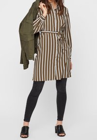 Abito a righe marrone e bianco, stretto in vita con una cintura, abbinato a legging scuri e sandali neri. Giacca verde drappeggiata.