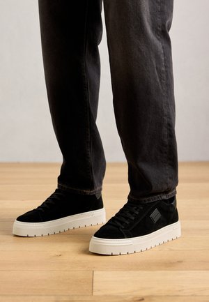 Person wearing black suede sneakers with white thick soles and dark jeans standing on a light wooden floor.