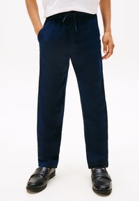 Copil purtând pantaloni bleumarin cu șnur și cu mâinile în buzunare, încălțat cu pantofi loafers negri, pe un fundal alb.