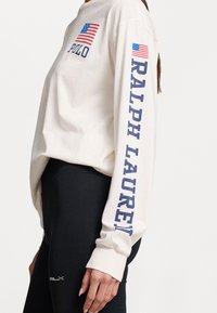 Ljusbeige långärmad skjorta med marinblå text "Polo" och "Ralph Lauren." Har en liten amerikansk flagga på vänster bröst och arm.