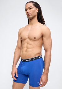 Hombre en forma con largas rastas, vistiendo calzoncillos bóxer azules de Nike, de pie frente a un fondo liso de color gris claro.