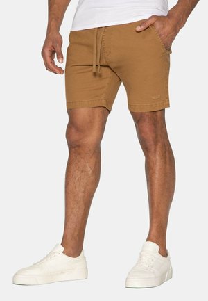 Pantaloni corti in cotone marrone con vita con cordino e tasche laterali, caratterizzati da una vestibilità aderente e bordi orlati. Abbinati a sneaker bianche.