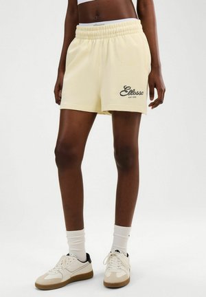 Osoba ubrana w jasnożółte szorty z elastycznym pasem i logo "Ellesse", w połączeniu z białymi skarpetkami i białymi trampkami z brązowymi podeszwami.