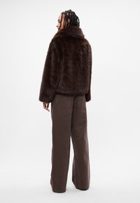 Casaco curto de pêlo sintético castanho com um colarinho alto, combinado com calças largas castanhas. A textura é fofa, acrescentando volume à silhueta.