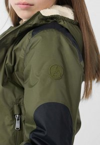 Giacca verde oliva con accenti neri, rivestimento interno morbido, chiusura con cerniera e toppa logo circolare sul braccio sinistro.