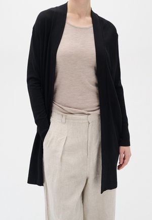 Osoba ubrana w jasnobeżową koszulę włożoną do wysokich beżowych spodni z plisami oraz długi czarny kardigan z otwartym przodem.