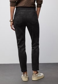 Pantalones de ante negros con un ajuste slim, que cuentan con dos bolsillos traseros y una textura suave. Llevados con zapatillas de color claro sobre una superficie gris.