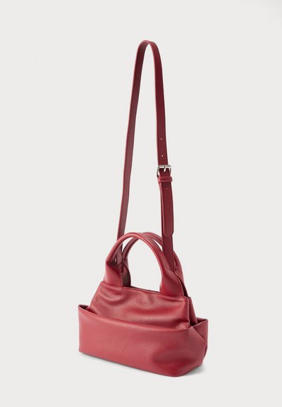 Μικρή κόκκινη δερμάτινη τσάντα χειρός με διπλές λαβές και ρυθμιζόμενο ιμάντα ώμου, που παρουσιάζεται όρθια σε απλό φόντο.