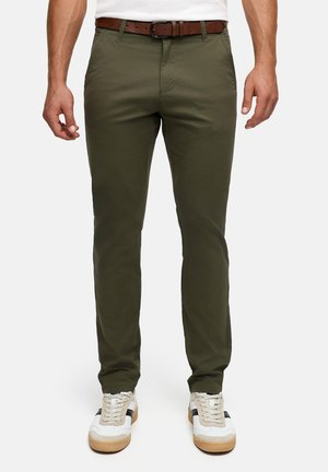 Homme portant un pantalon ajusté vert olive avec une ceinture en cuir marron et des baskets blanches à semelles en gomme, debout devant un fond uni.
