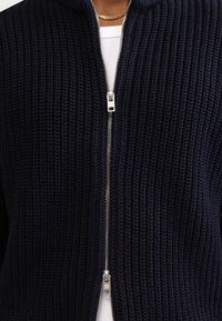 Marineblaue Strickjacke mit Reißverschluss und strukturiertem Muster, ausgestattet mit einem silbernen Reißverschluss und einer Kapuze. Hergestellt aus dicker, weicher Material für Wärme.