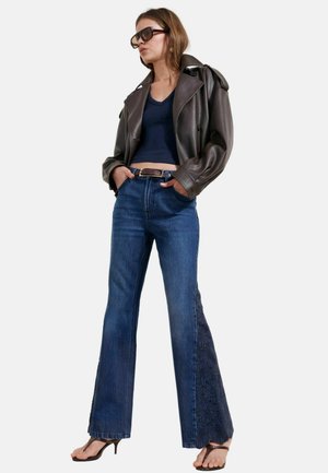 Kvinde iført mørkebrun læderjakke, navy crop top, blå viddebukser med teksturerede sidelommer, brun bælte, solbriller og brune hælede sandaler.